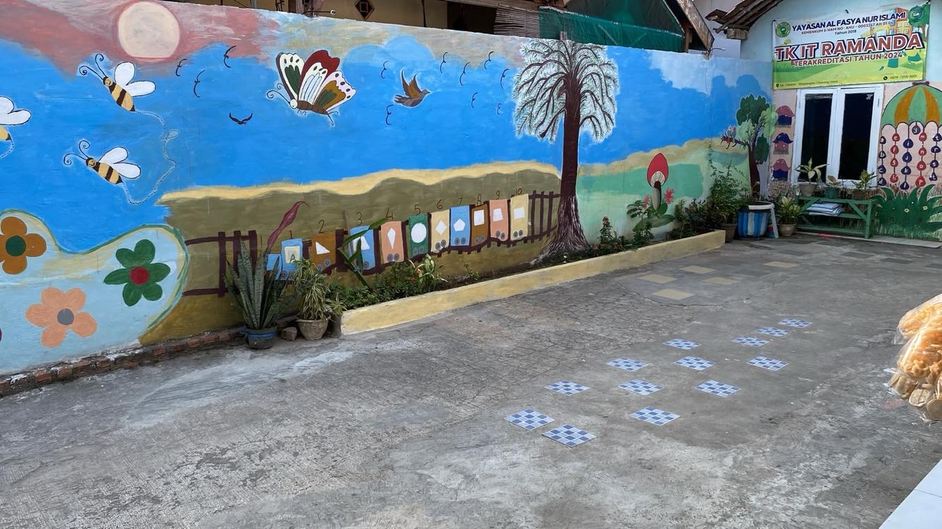 Halaman TK Islam Ramanda dengan mural langit biru, kupu-kupu, lebah, dan pohon, serta plang Yayasan Al Fasya Nur Islami di depan kelas
